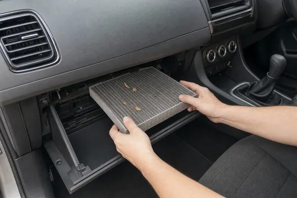 Hands sliding a grey, dusty cabin filter with dry leaves out of its slot